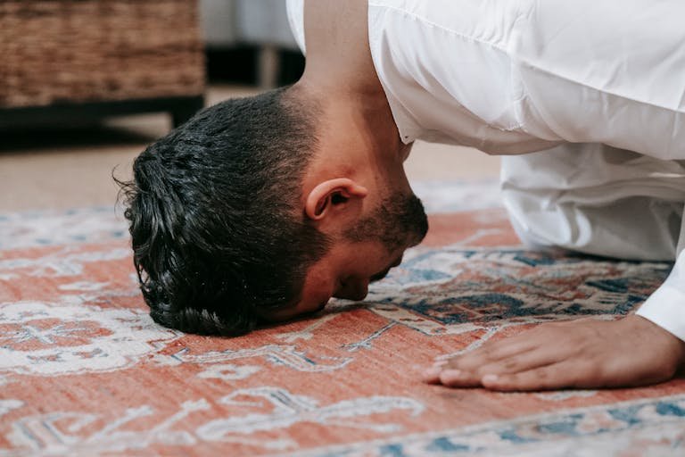 A man in a white thobe performs salah, kneeling on a patterned prayer rug indoors.