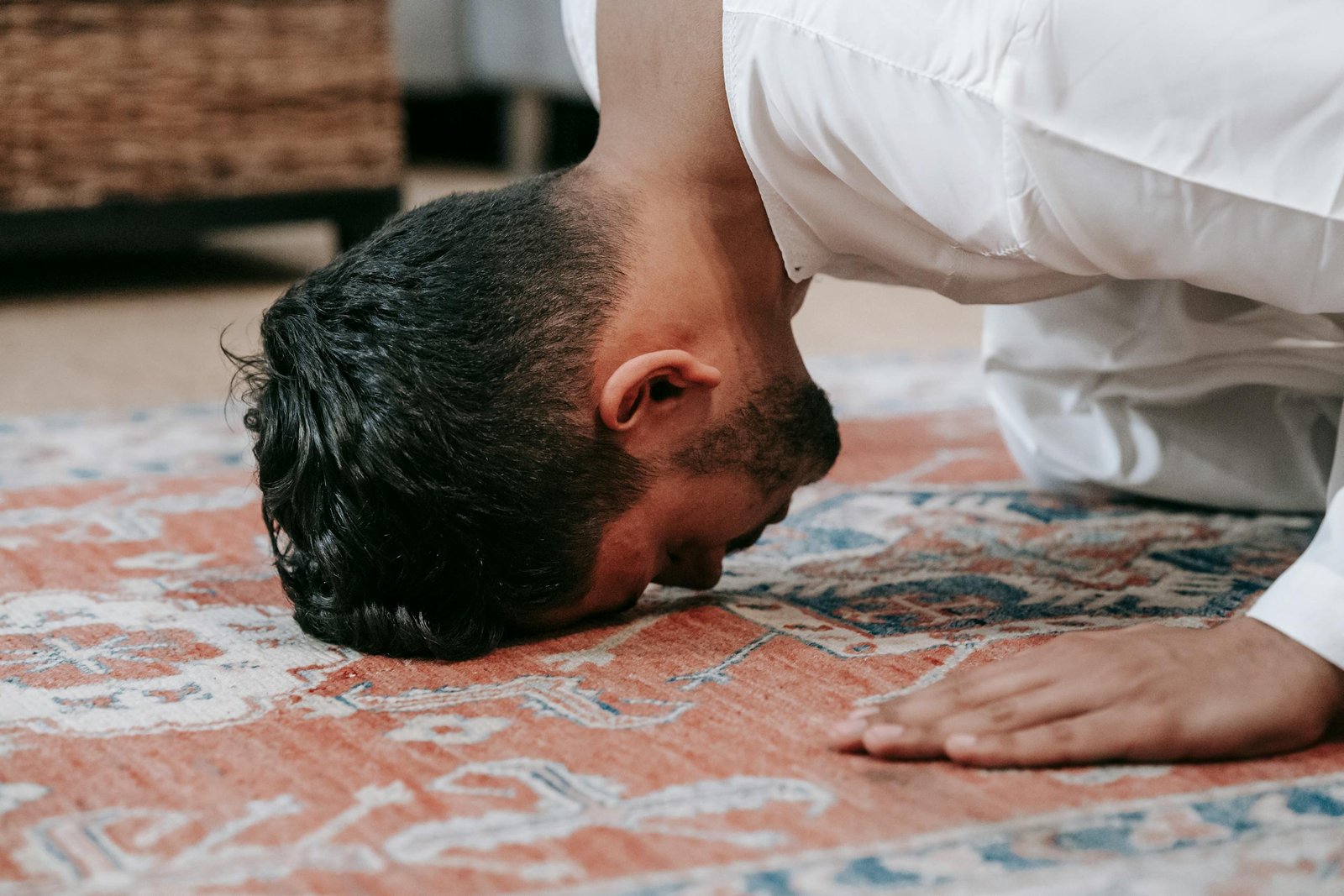 A man in a white thobe performs salah, kneeling on a patterned prayer rug indoors.